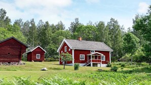 Une maison en bois rouge, dotée d’un porche et d’un petit abri de jardin rouge situé à proximité, le tout entouré d’une végétation luxuriante et d’arbres.