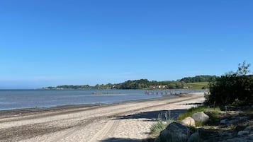 A sandy beach with a clear blue sky, a rocky shoreline, and a distant view of a small town with houses and greenery.