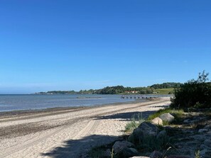 A sandy beach with a clear blue sky, a rocky shoreline, and a distant view of a small town with houses and greenery. - 4 Star Holiday Home in Loddekopinge (Loddekopinge)