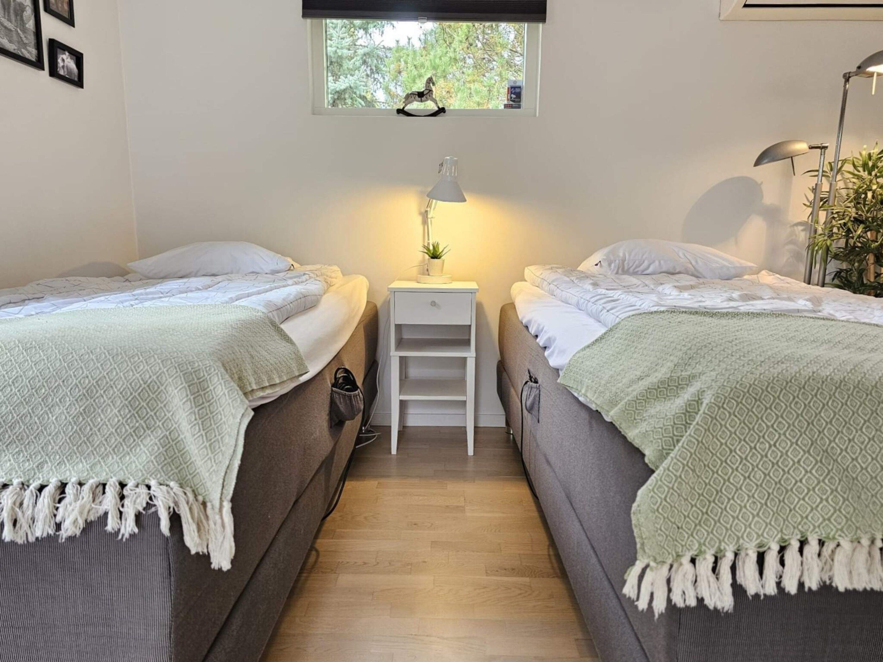 A room with two beds, a small white nightstand with a lamp, a window with a view of trees, and a plant on the right.