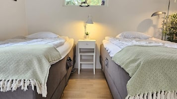 A room with two beds, a small white nightstand with a lamp, a window with a view of trees, and a plant on the right.