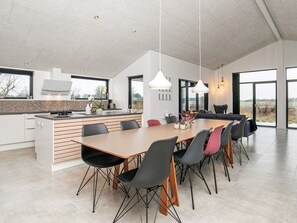 A modern kitchen with a large island, dining area with chairs, pendant lights, and large windows offering a view of the outdoors.