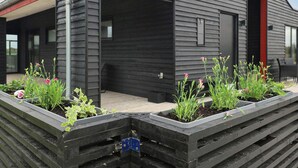 Un bâtiment moderne à l’extérieur sombre, doté d’un espace extérieur couvert et de jardinières surélevées, ainsi que d’une porte rouge.