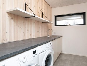 A modern kitchen with a built-in washing machine, wooden wall paneling, a window, and a metal shelf. - 5 Star Holiday Home in Lokken (Lokken)