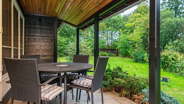 Eine überdachte Terrasse mit Holzboden, einem runden Tisch und vier Korbstühlen mit Kissen.