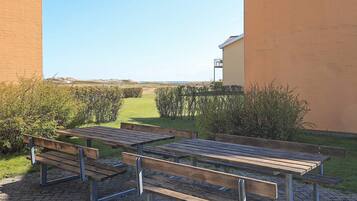 An outdoor seating area with picnic tables and benches, surrounded by a hedge and a building.