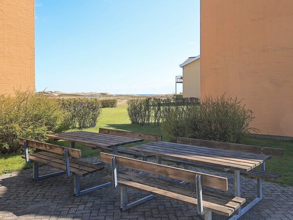 An outdoor seating area with picnic tables and benches, surrounded by a hedge and a building.