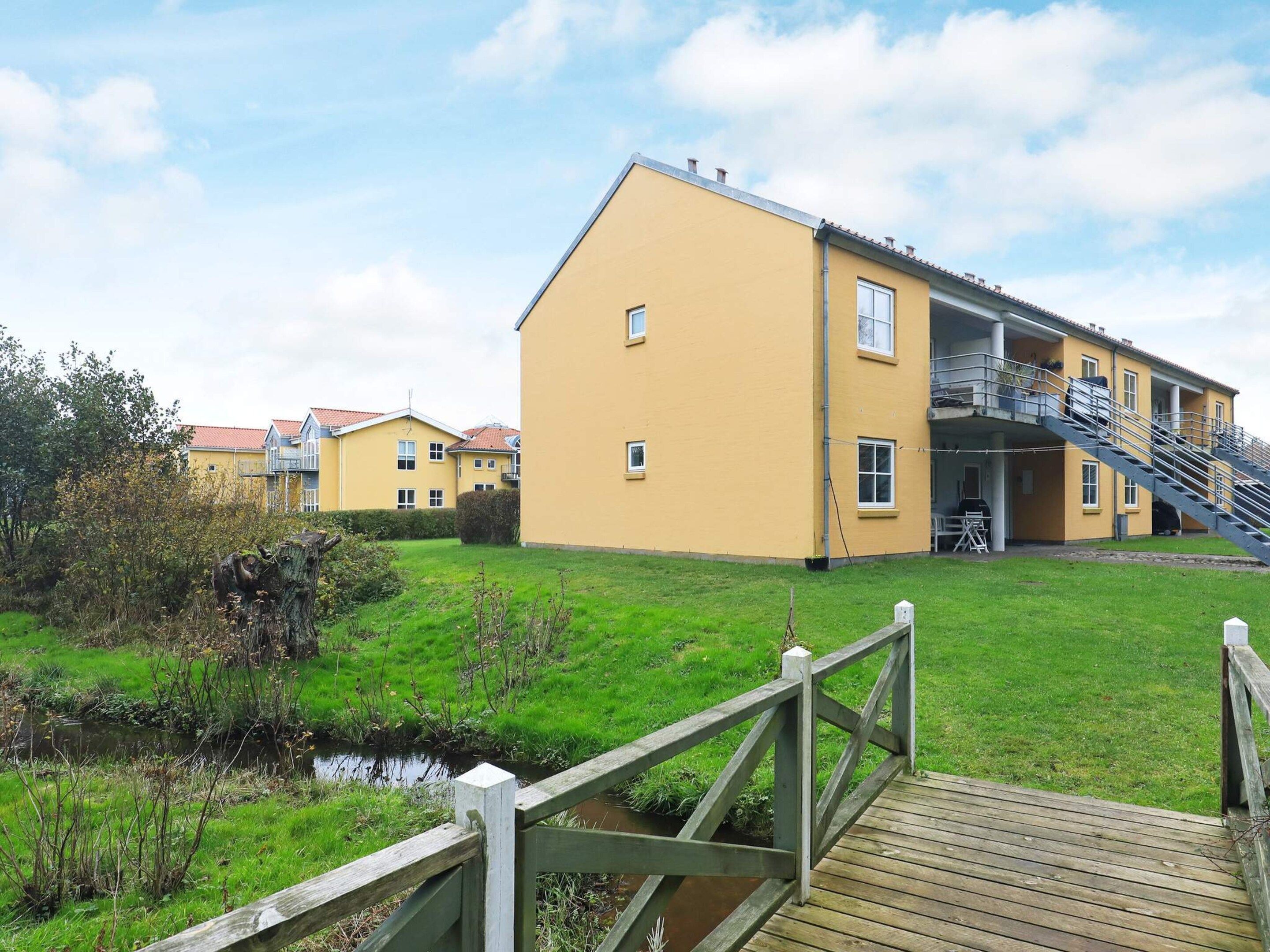 A yellow building with a staircase leading to a balcony, a wooden deck, and a small stream in the foreground.