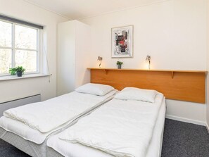 A room with two single beds, a window with blinds, a wall-mounted shelf with a plant, and a framed picture above the beds.