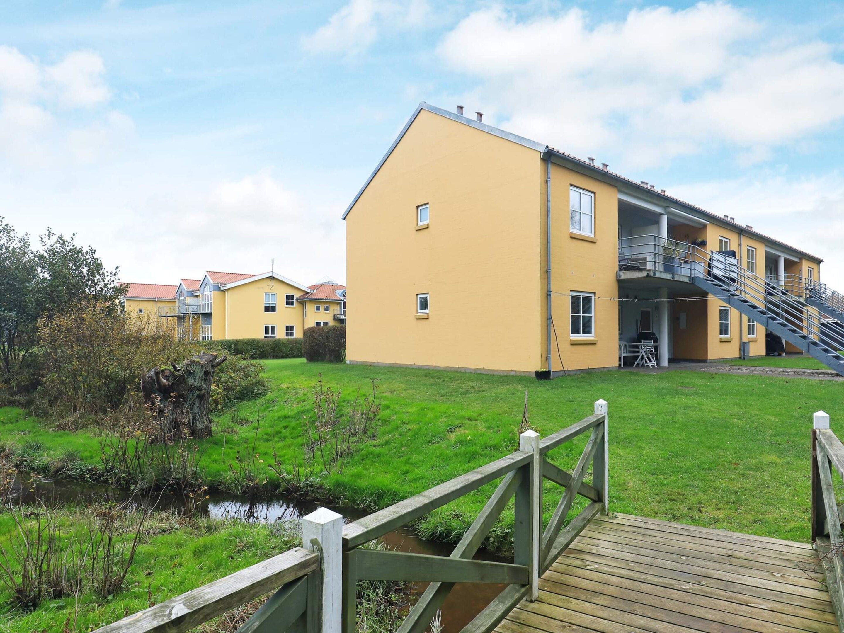 A yellow building with a staircase leading to a balcony, a wooden deck, and a small stream in the foreground.