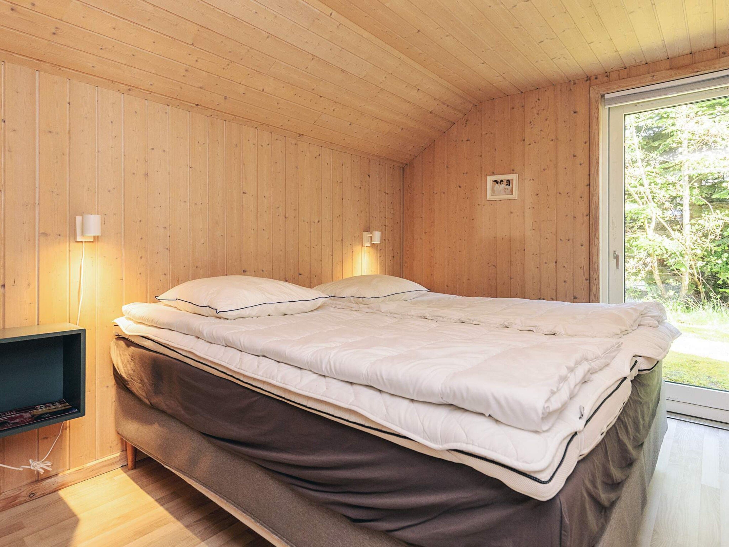Egy fa borítással ellátott szoba, amelyben egy ágy, két falra szerelt lámpa és egy ablak található, ahonnan zöld tájra nyílik kilátás.