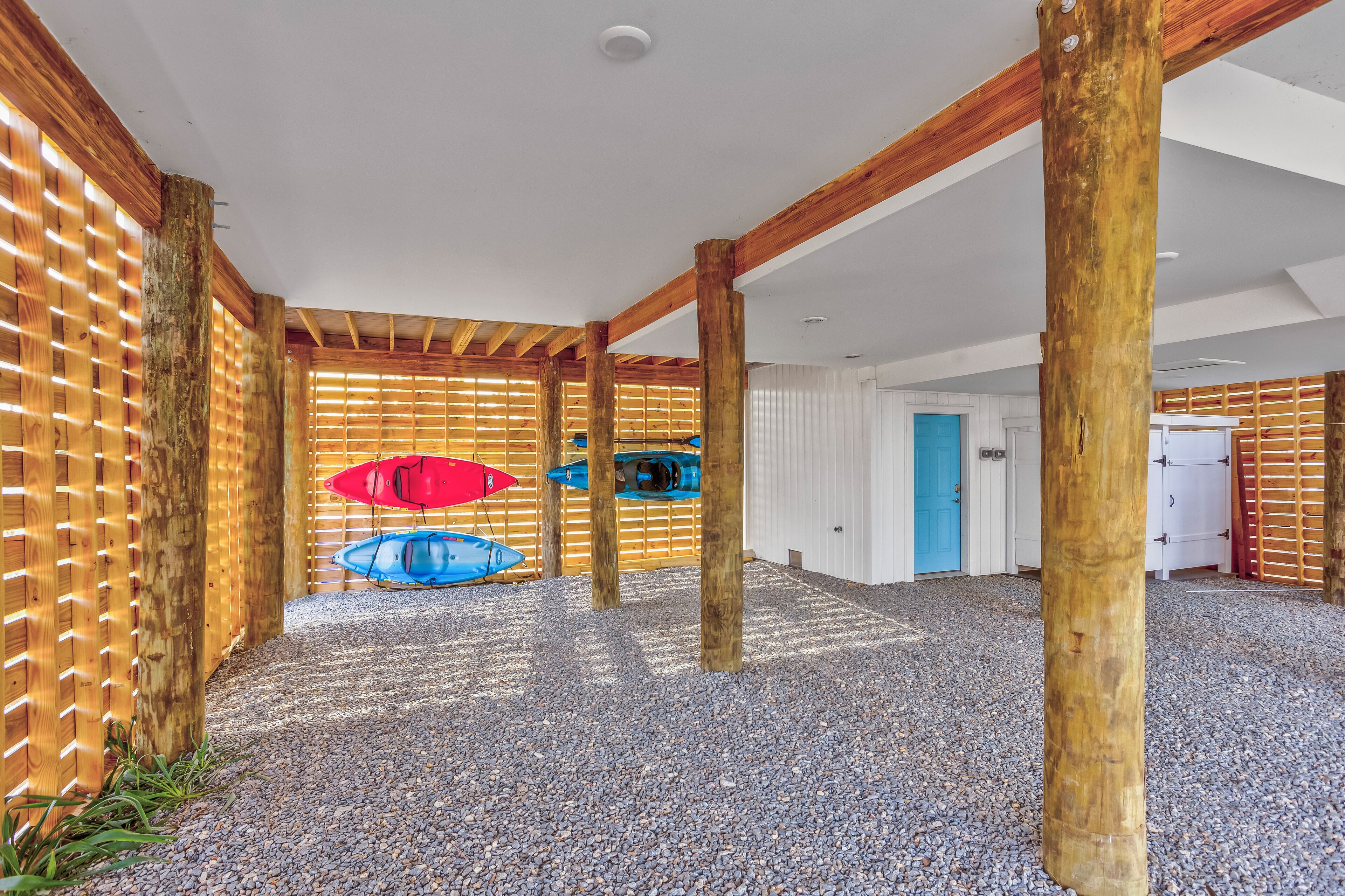 Un espacio al aire libre cubierto con vigas de madera y suelo de grava, con dos kayaks y una sombrilla roja.