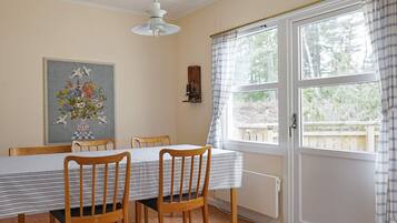 A dining area with a table set for four, wooden chairs, a hanging light fixture, a window with curtains, and a wall-mounted shelf with items.