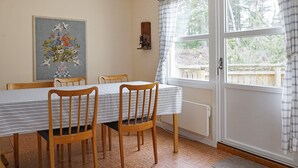 A dining area with a table set for four, wooden chairs, a hanging light fixture, a window with curtains, and a wall-mounted shelf with items.