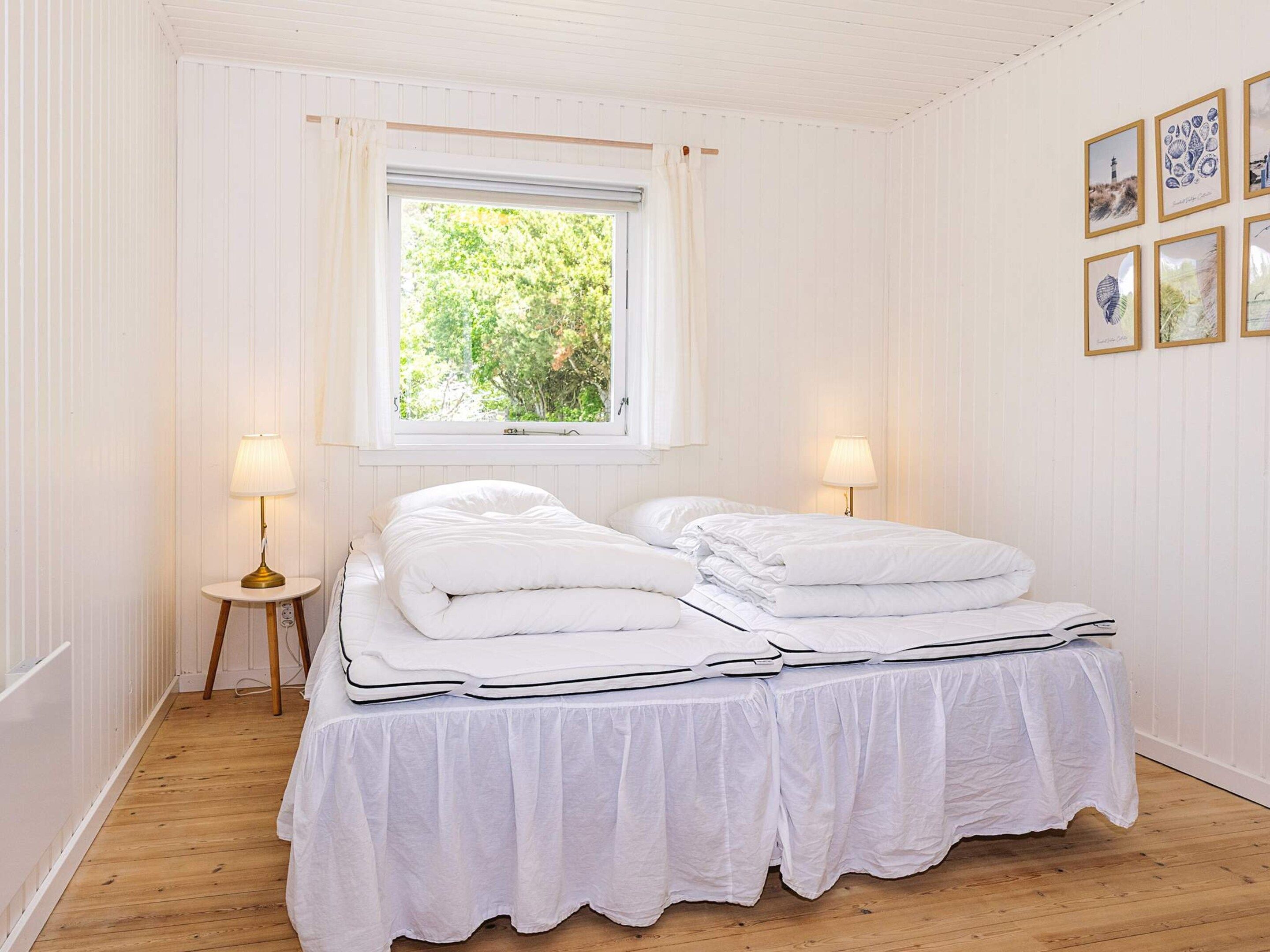 A room with a bed, two bedside lamps, a window with a view of greenery, and framed pictures on the wall.