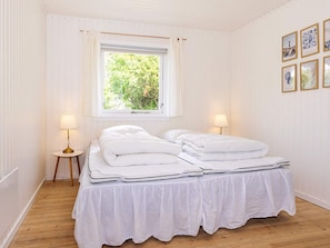 A room with a bed, two bedside lamps, a window with a view of greenery, and framed pictures on the wall.