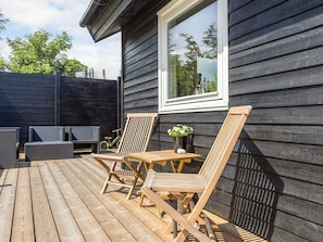 A wooden deck with outdoor furniture, including chairs and a table, adjacent to a building with dark siding and a window.
