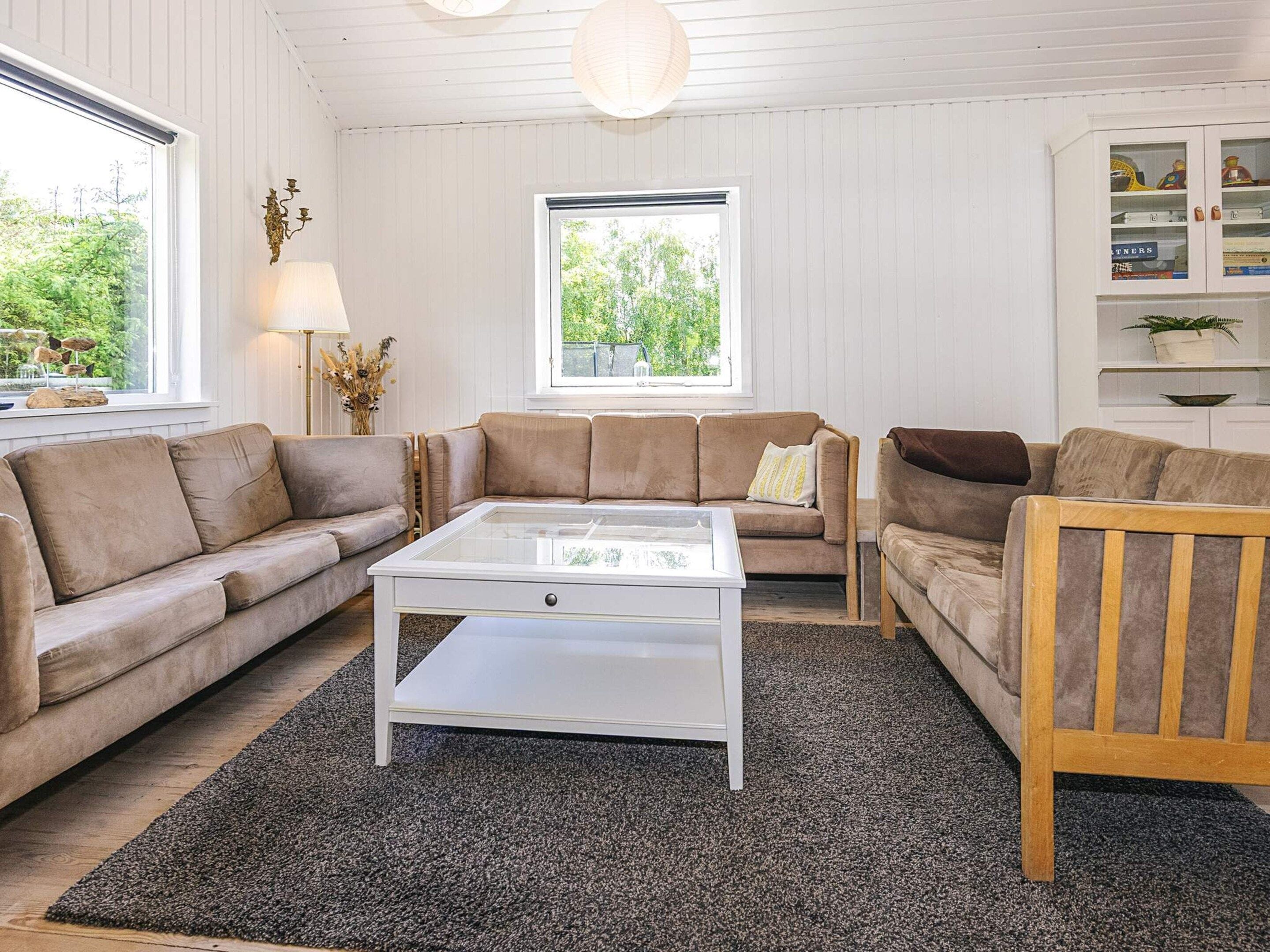 Un salon avec un canapé beige, une table basse blanche et une chaise en bois. Deux fenêtres laissent entrer la lumière naturelle et offrent une vue sur la verdure à l’extérieur.