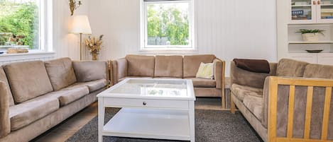 A living room with a beige sofa, a white coffee table, and a wooden chair. There are two windows providing natural light and a view of greenery outside.