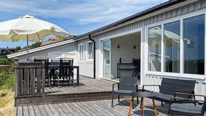 Een houten terras met een tafel en stoelen, een wit gebouw met grote ramen, en een witte parasol.