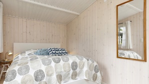 Een slaapkamer met een bed, een nachtkastje, een lamp, een spiegel en een rieten mand met boeken.