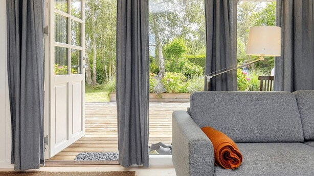 Een moderne woonkamer met een grijze bank, een opgerolde oranje deken, en uitzicht op een tuin door een glazen deur.