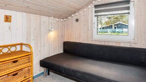 Une pièce avec un plafond en bois, un canapé noir, une commode en osier, un luminaire fixé au mur et une fenêtre donnant sur l’extérieur.