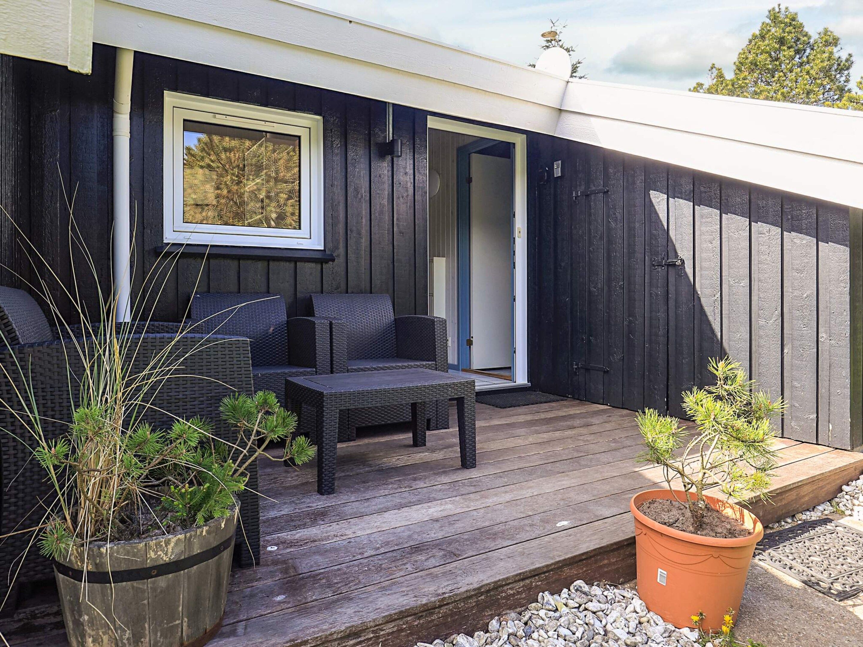 Una terraza de madera con mobiliario exterior, una planta en maceta y una puerta que da al interior.