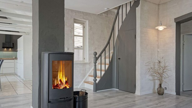 Un salon moderne avec une cheminée centrale, un escalier et une cuisine en arrière-plan.