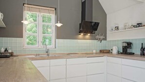 Une cuisine moderne dotée d’un grand îlot central, de meubles blancs, d’une fenêtre avec stores et d’une machine à café posée sur le plan de travail.
