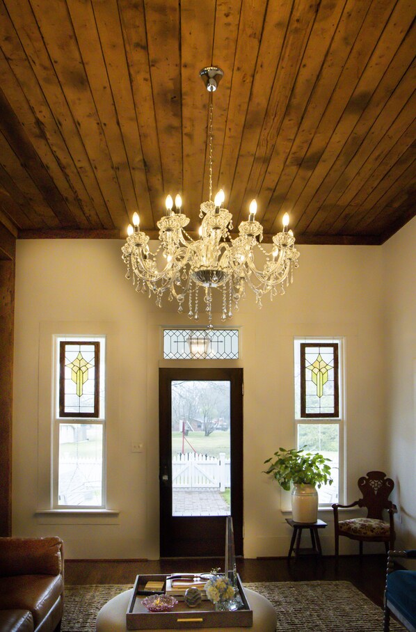 A living room with a wooden ceiling, a chandelier, two stained glass windows, a door, a potted plant, and a brown leather sofa. - The Cottage on Maple (Murfreesboro)