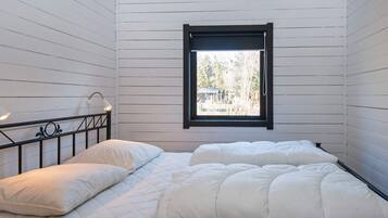 A single bed with a white duvet and pillows in a room with white wooden walls and a window with a black frame.