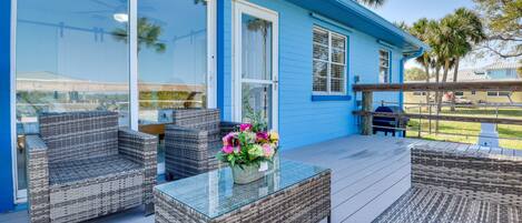 A blue house with a covered patio area featuring wicker furniture and a glass-top table with a flower arrangement.