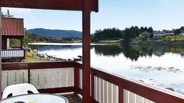 Een houten terras met een glazen tafel en stoelen, met uitzicht op een rustig meer met huizen op de achtergrond.