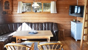 Een gezellig cabin interieur met een houten plafond, een leren bank, een eettafel, rieten stoelen, een televisie en een dressoir.