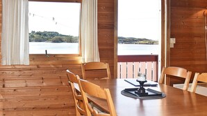 Een houten eetkamer met een tafel en stoelen, grote ramen met gordijnen en uitzicht op een waterpartij en heuvels.