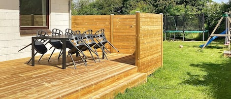 Une terrasse en bois avec une table et des chaises, une zone délimitée par une clôture et, au fond, une aire de jeux.