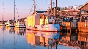 Escena frente al agua con un barco cubierto amarrado en un muelle, un edificio con un restaurante llamado 'SHANGOOR' al fondo, y agua tranquila que refleja los barcos y el edificio.