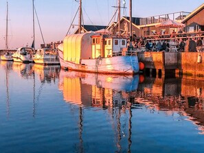 A waterfront scene with a covered boat docked at a pier, a building with a restaurant named 'SHANGOOR' in the background, and calm water reflecting the boats and building. - 10 Person Holiday Home in Vinderup-by Traum (Vinderup)
