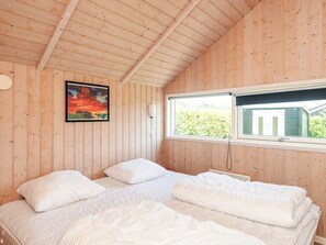 A room with wooden walls and a sloped ceiling, featuring a bed with white bedding, a window with a view of greenery, and a painting on the wall.