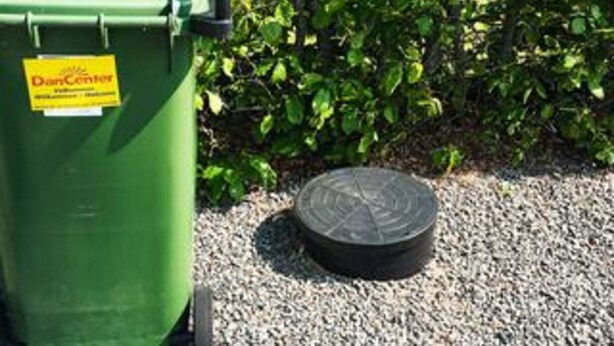 A green dumpster with a yellow label and a black circular object on gravel. - 5 Star Holiday Home in Ansager (Ansager)