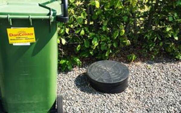 A green dumpster with a yellow label and a black circular object on gravel. - 5 Star Holiday Home in Ansager (Ansager)