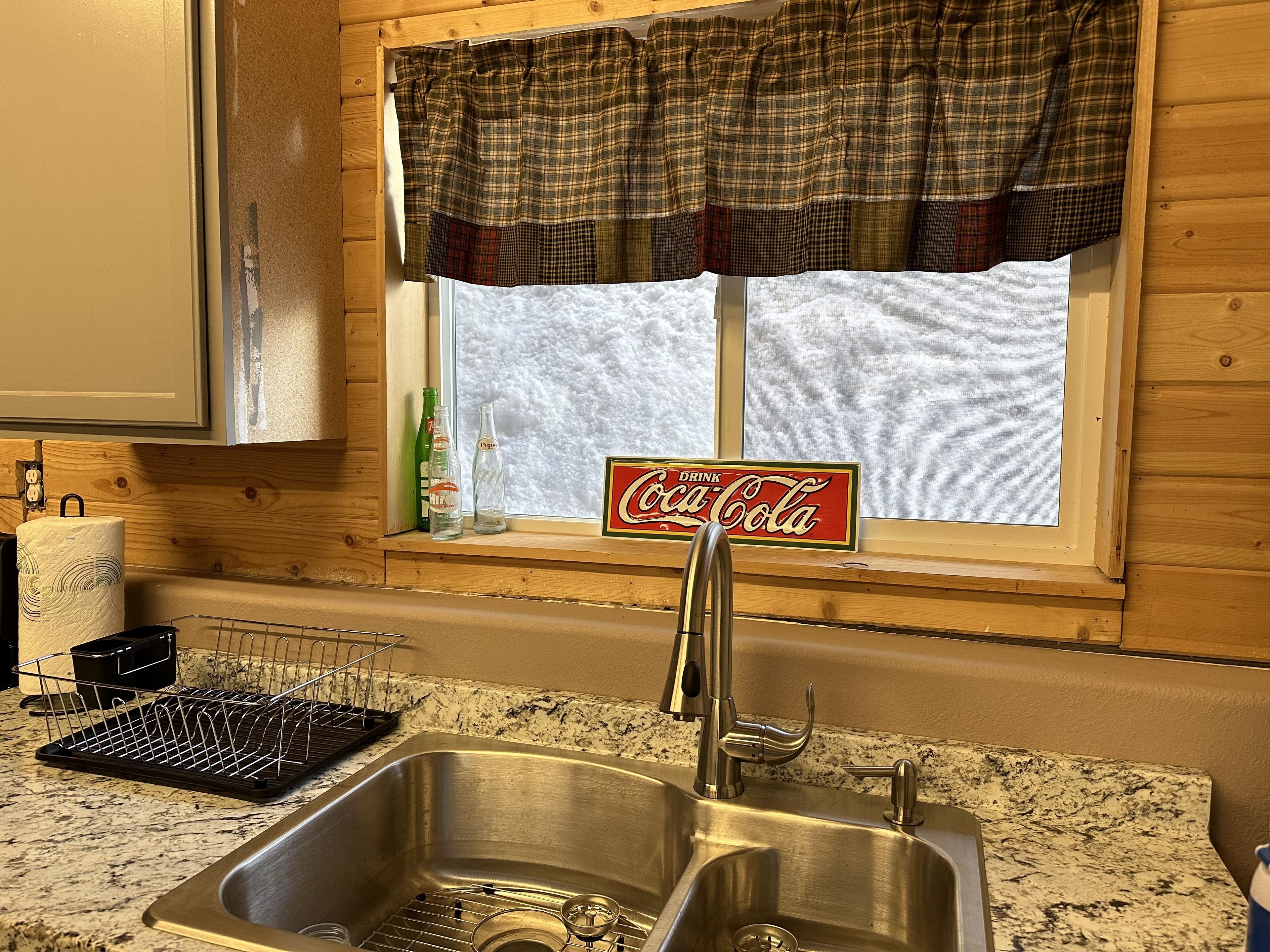 A kitchen with a stainless steel sink, a window with a plaid valance, and a Coca-Cola sign.