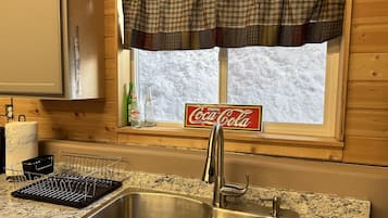 A kitchen with a stainless steel sink, a window with a plaid valance, and a Coca-Cola sign.