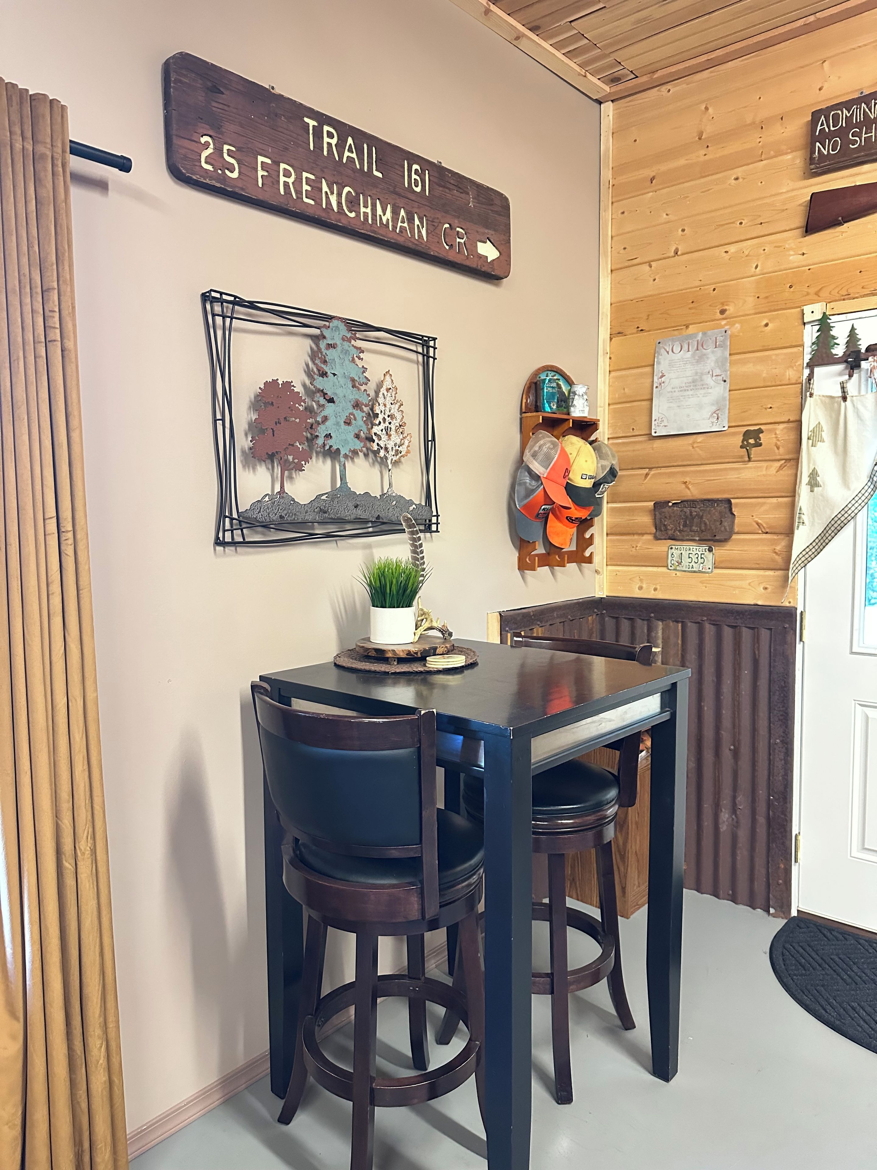A wooden interior with a table and chairs, a sign indicating 'Trail 161 25 Frenchman CR', and various wall decorations including a notice board and a helmet.
