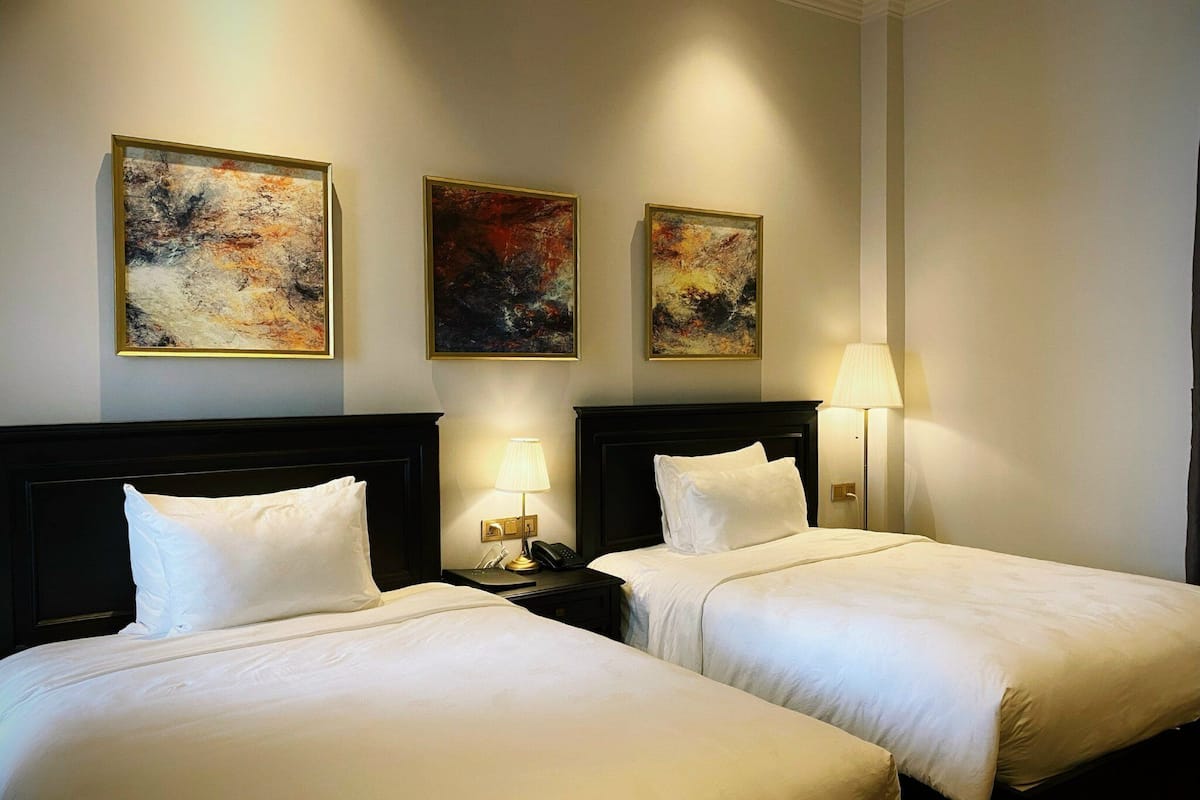 A hotel room with two beds, a nightstand, a lamp, and three abstract paintings on the wall.