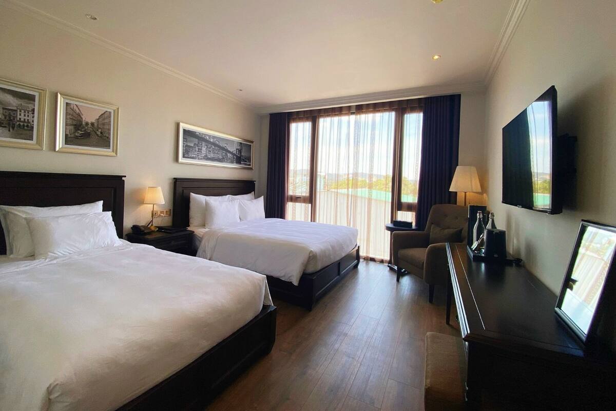 A hotel room with two beds, a desk with a TV, and a view of the outdoors.