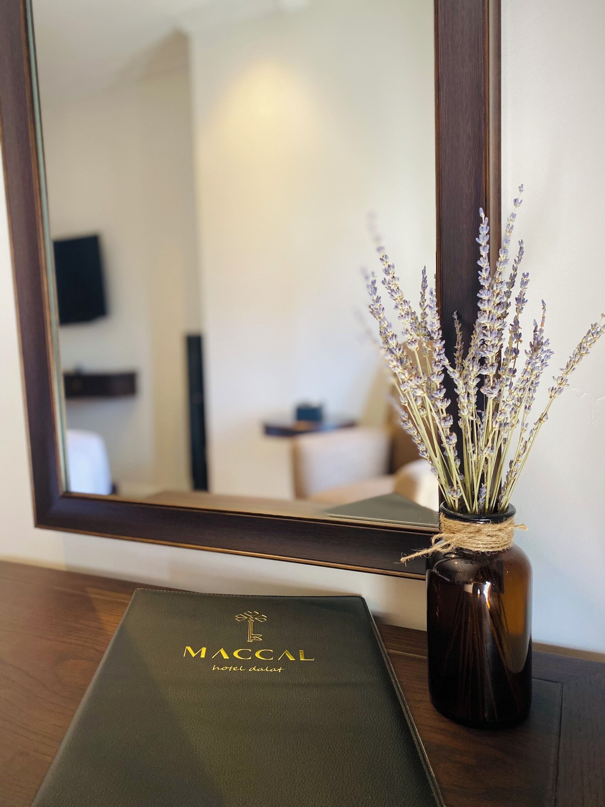 A dark brown vase with lavender flowers on a wooden surface, a mirror reflecting a room with a sofa and a small table, and a green booklet with 'MACCAL hotel dalat' text.