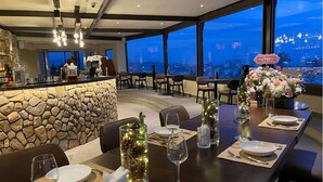 Restaurante com parede de pedra, mesas postas para refeições e vista da cidade ao entardecer.