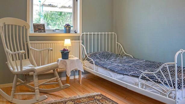 Une chambre avec un lit simple, un fauteuil à bascule, une petite table avec une lampe et une fenêtre donnant sur un paysage verdoyant.